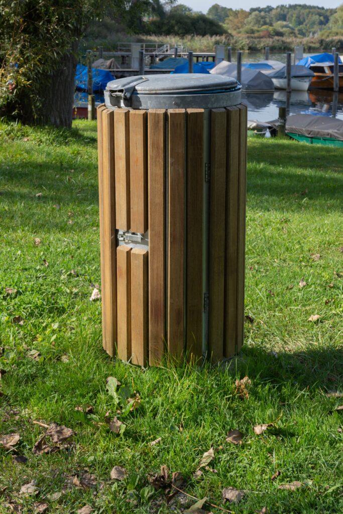 Runder Mülleimer mit Holzverkleidung und Metalldeckel, der auf einer Wiese in einem Park neben einem See mit Booten steht.