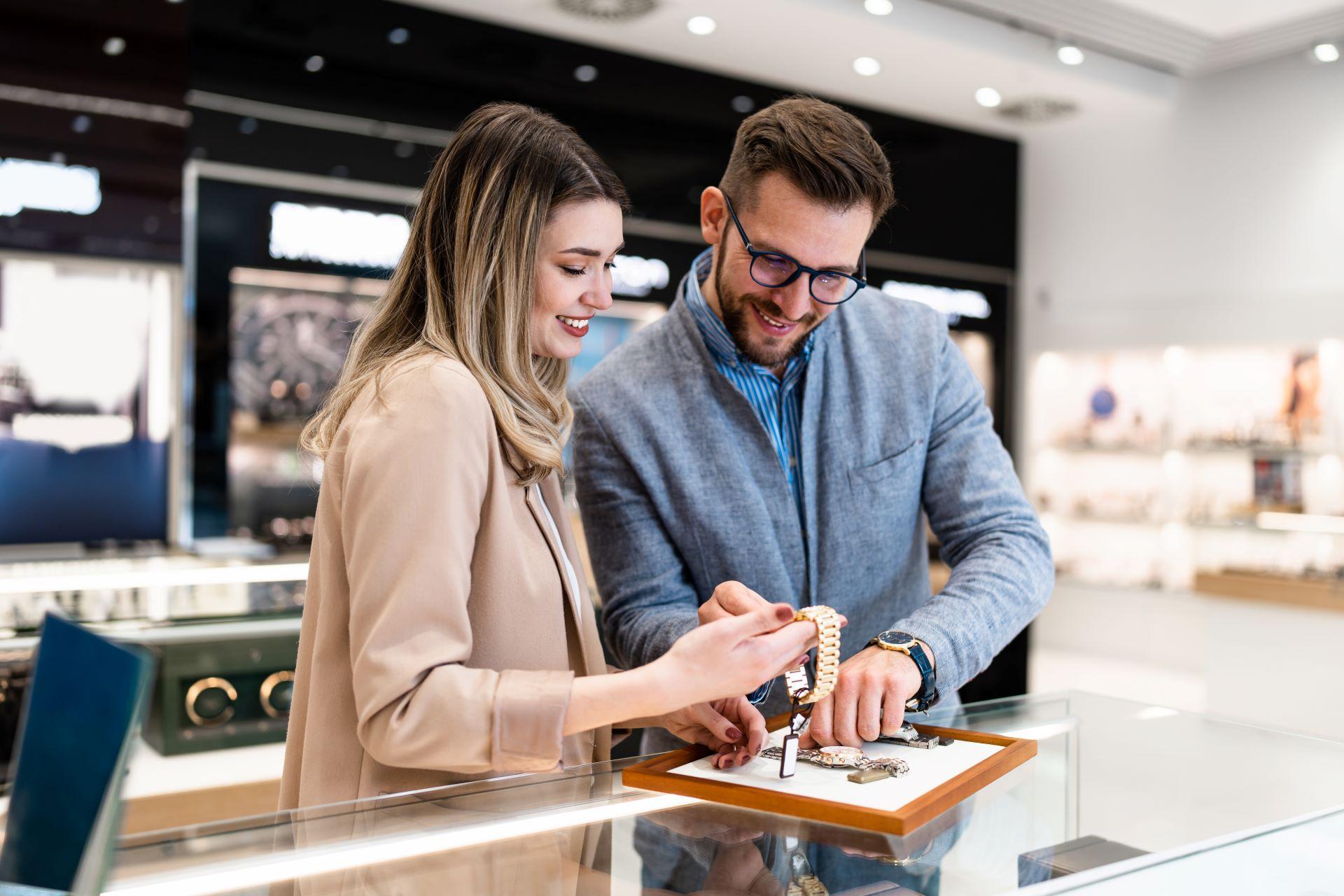 Ein lächelndes Paar begutachtet gemeinsam eine goldene Armbanduhr in einem modernen Juweliergeschäft, während der Verkäufer den Schmuck präsentiert.