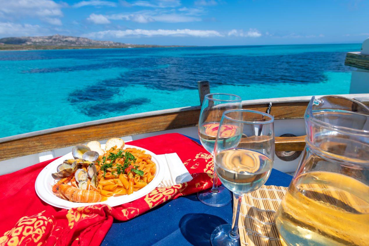 Teller mit Meeresfrüchte-Pasta und Weißwein auf einem Boot, türkisblaues Wasser in Sardinien im Hintergrund 